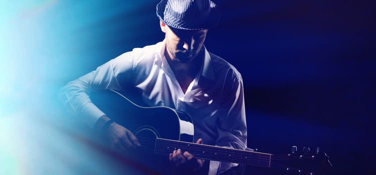 a musician sits on a dark stage with one spotlight holding a guitar and looking down